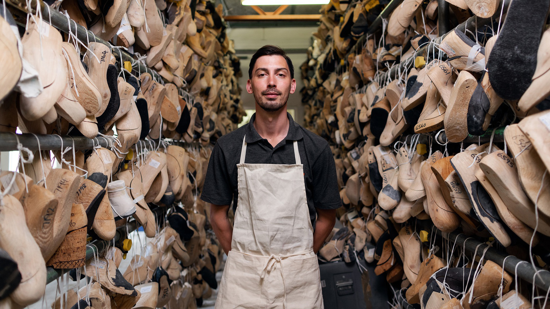 Handwerker in Leinenschürze steht zwischen dicht gehängten Schuhleisten in einem Manufaktur-Lager