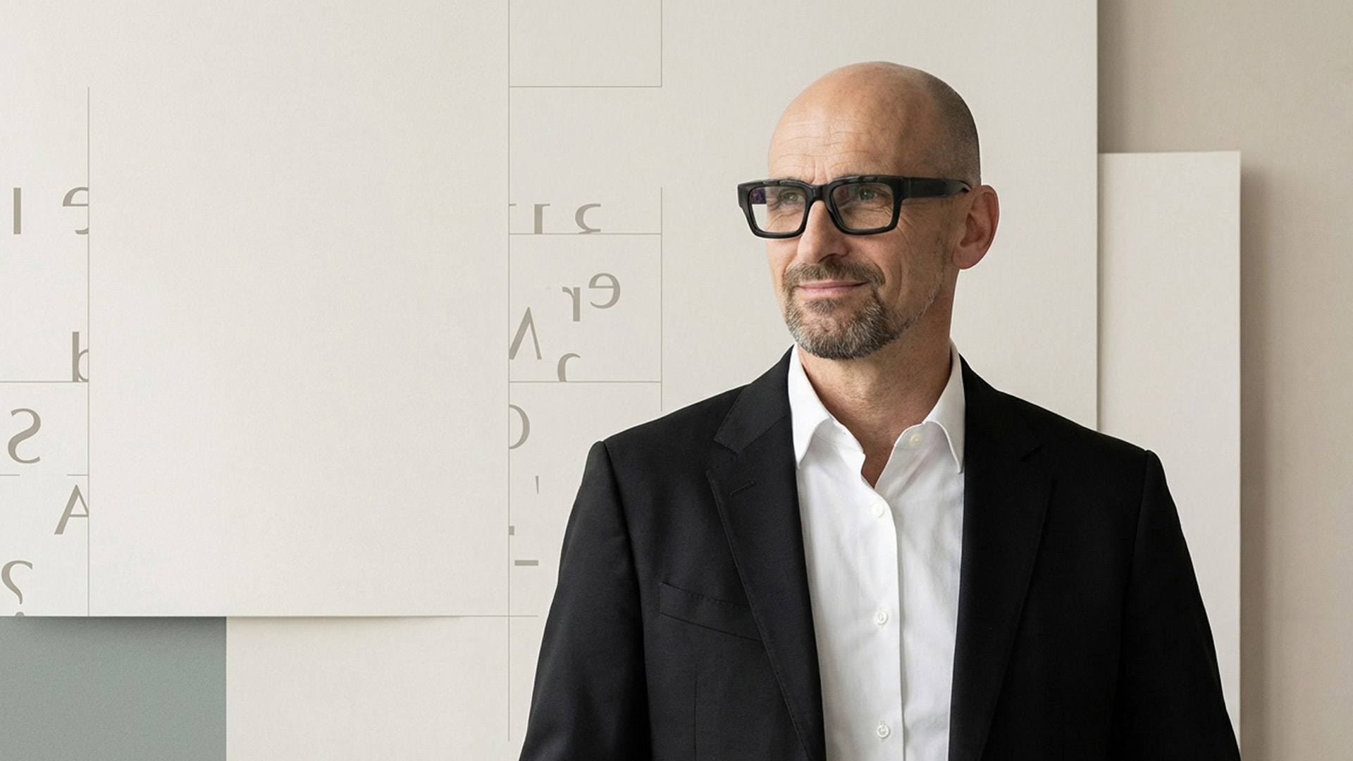 Porträt von Stephan Göckler mit Brille und dunklem Sakko vor einer Wand mit großformatiger, unregelmäßig angeordneter Typografie-Collage.