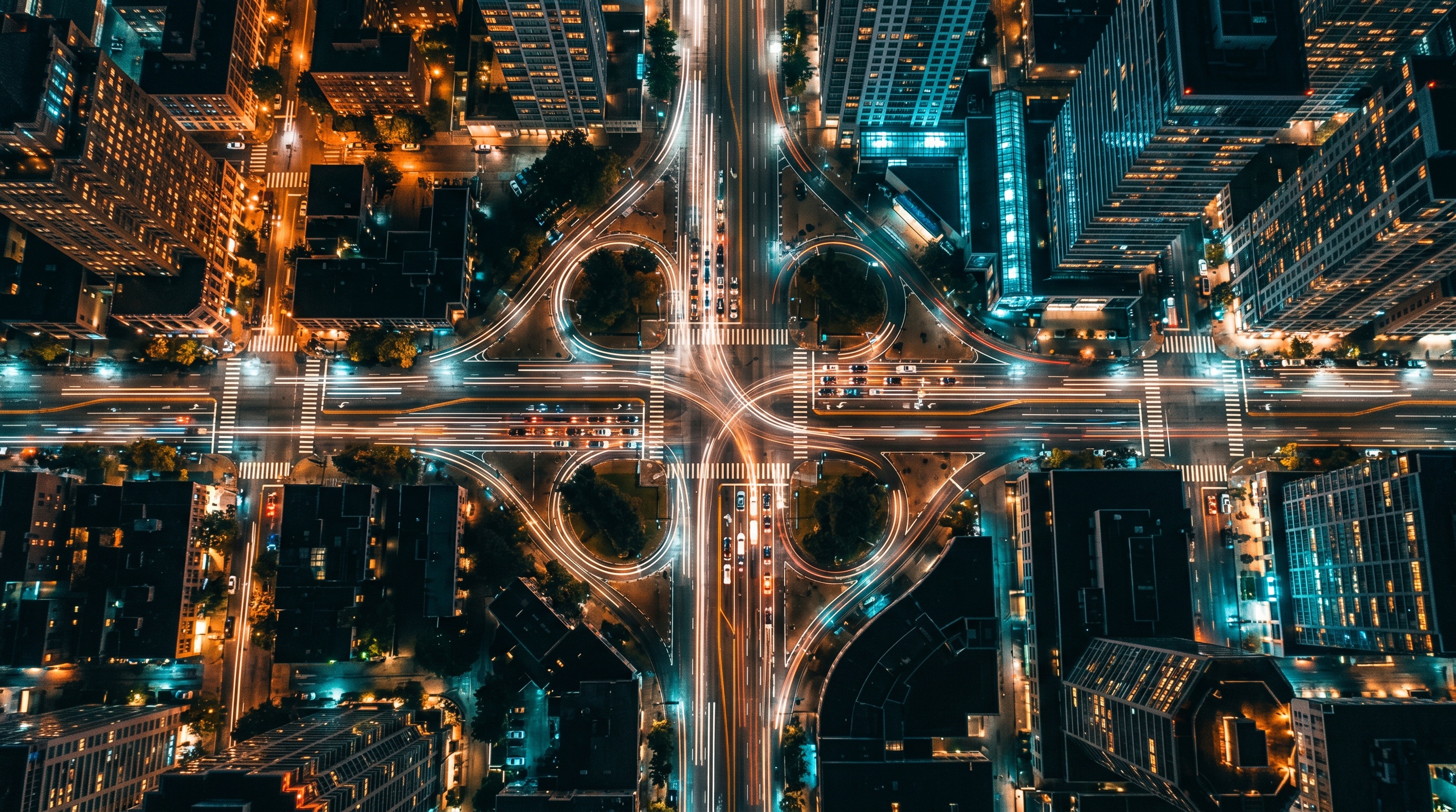 Luftaufnahme einer nächtlichen Straßenkreuzung mit mehreren sich kreuzenden Fahrspuren und leuchtenden Lichtspuren des Verkehrs, umgeben von Hochhäusern, als Sinnbild für viele zusammenlaufende Wege, die erst aus der Vogelperspektive einen Zusammenhang ergeben.